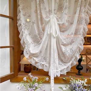 Op de afbeelding: Witte, transparante gordijn met bloemen- en vlindermotieven. Het gordijn heeft een gerimpelde rand en is vastgebonden met een bijpassende stoffen band. Het gordijn hangt voor een raam.