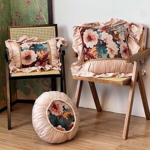 May include: Two wooden chairs with woven seats and backs, each adorned with a floral patterned pillow. A round pillow with the same floral pattern sits on the floor. The pillows have a pink ruffled trim. The floral pattern includes pink, teal, and burgundy.
