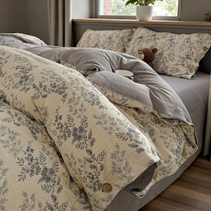May include: A bed dressed with a floral duvet cover and pillowcases. The duvet cover features a cream backdrop with a grey floral pattern. The bed also includes a grey sheet and a grey comforter. Wooden buttons are visible on the duvet cover.