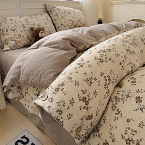 Peut inclure: Une parure de lit composée d'une housse de couette et de taies d'oreiller beiges à motif floral marron foncé avec des boutons ronds visibles. L'ensemble comprend une section en tissu marron clair uni et un petit ours en peluche marron.