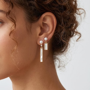 Puede incluir: Un par de pendientes dorados asimétricos. Uno tiene un pequeño perno de perla blanca con un colgante rectangular corto con incrustación blanca. El otro tiene un perno de perla, una pequeña piedra transparente y un colgante rectangular largo.
