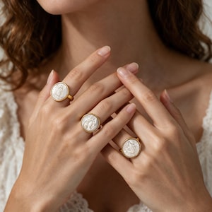May include: Three gold-toned rings featuring circular white mother-of-pearl inlays. Each ring displays a detailed embossed religious figure centered on the iridescent surface, set within a polished gold metal band and circular frame.