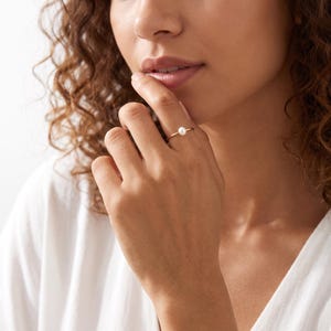 May include: A thin gold band ring featuring a single small, round, white pearl centred on the top. The ring is worn on the ring finger, showcasing the delicate metal wire construction and the smooth, lustrous surface of the pearl gemstone.