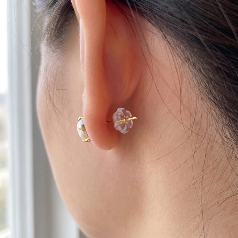 May include: A gold stud earring featuring a round white stone set in a gold frame, secured at the back with a translucent, flower-shaped silicone earring back. The earring is positioned through an earlobe, showing the front stone and back closure.