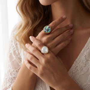 May include: Two gold-toned rings featuring irregular, organic-shaped settings. One ring displays a polished, iridescent abalone shell inlay with swirling green and purple hues, while the other ring features a smooth, opaque white stone inlay.