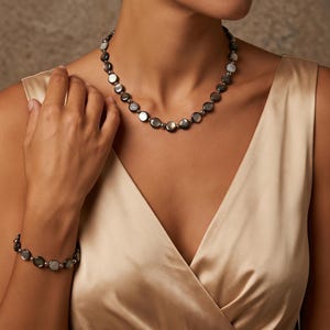Könnte beinhalten: Ein Halsketten- und Armbandset bestehend aus runden, irisierenden dunkelgrauen Muschelperlen, die mit kleinen, polierten silberfarbenen Metallkugeln abwechseln. Der Schmuck wird zu einem hellen, champagnerfarbenen Satinkleid getragen.
