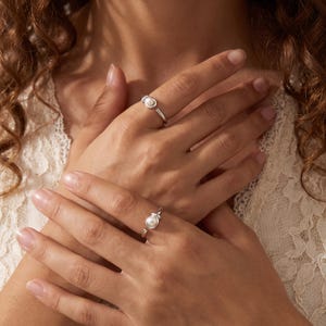 Puede incluir: Dos anillos abiertos de tono plateado, cada uno con una pequeña perla blanca engastada en un extremo. Los anillos tienen un acabado metálico liso y pulido, con un diseño de banda simple y ajustable que permanece abierto en la parte frontal.