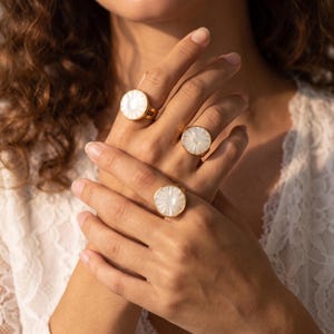 May include: Three gold-toned rings featuring circular, white, iridescent, shell-like inlays with a radial, sunburst-style pattern. Each ring has a thin, polished gold band and is worn on the fingers, showcasing the reflective, pearlescent surface.