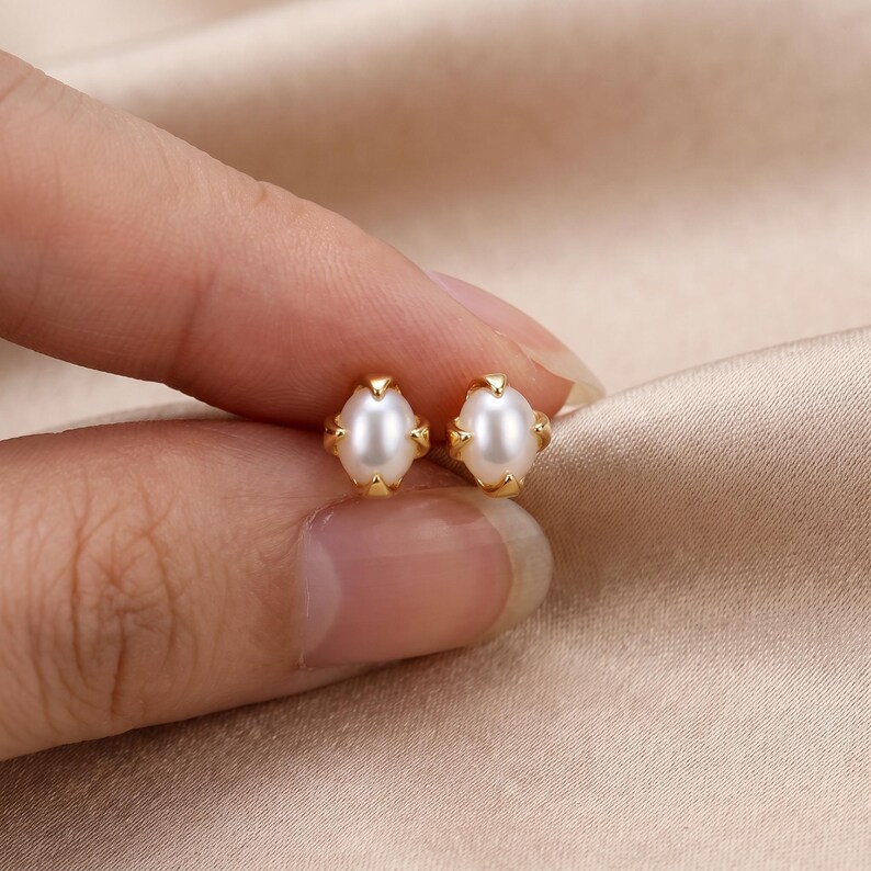 May include: A pair of stud earrings featuring oval-shaped white pearls set in gold-toned metal prongs. Each earring has a smooth, lustrous finish and a secure four-prong setting that holds the pearl firmly in place, creating a classic jewelry piece.