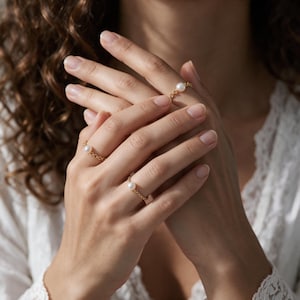 Puede incluir: Tres delicados anillos de cadena dorada, cada uno con una pequeña perla blanca. Los anillos se llevan en los dedos, mostrando una fina construcción de eslabones metálicos con un acabado pulido y un acento de perla blanca redonda.