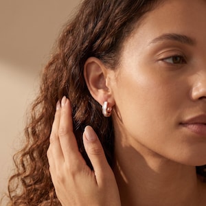 May include: A small hoop earring featuring a metallic rose gold-toned band with a central inlay of iridescent white mother-of-pearl. The earring has a circular shape and is worn on the earlobe, showcasing a polished finish and a smooth texture.