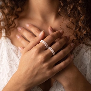Könnte beinhalten: Zwei Ringe aus kleinen, runden, weißen Perlen. Jeder Ring besteht aus einem einzelnen Perlenband, das den Finger umschließt. Die Perlen sind in Größe und Farbe einheitlich und bilden ein zartes, durchgehendes kreisförmiges Design.