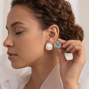 May include: A pair of stud earrings featuring irregular, rounded shapes with gold-toned metal edges. One earring displays a smooth, iridescent white shell inlay, while the other showcases a vibrant, multi-colored abalone shell pattern.