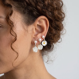 Op de afbeelding: Drie individuele oorbellen met witte bloemmotieven. Eén oorbel is een witte margriet met een geel hart, terwijl de andere twee witte uitgesneden rozenvormen zijn. Elke oorbel is geaccentueerd met een kleine witte parel en een heldere steen.