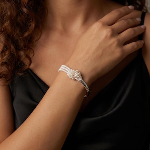 May include: A silver-toned cuff bracelet featuring three parallel strands of small white pearls that converge into a central cluster knot. The bracelet has a smooth, polished metallic finish and a structured, open-ended design that rests on the wrist.