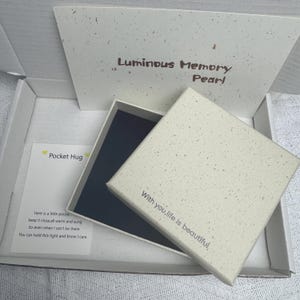 May include: A square off-white gift box with a speckled texture, featuring the text With you, life is beautiful on the lid. Inside the open box is a dark blue lining. A card labeled Pocket Hug with a poem is included in the white shipping box.