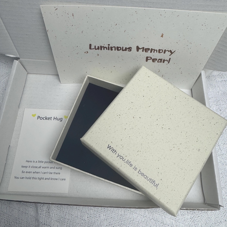 May include: A square off-white gift box with a speckled texture, featuring the text With you, life is beautiful on the lid. Inside the open box is a dark blue lining. A card labeled Pocket Hug with a poem is included in the white shipping box.