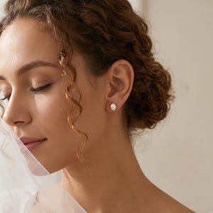 May include: A small, oval-shaped stud earring featuring a central white pearl set within a gold-toned metal frame. The earring is secured to the earlobe, showcasing a polished finish and a classic, symmetrical design that highlights the pearl.