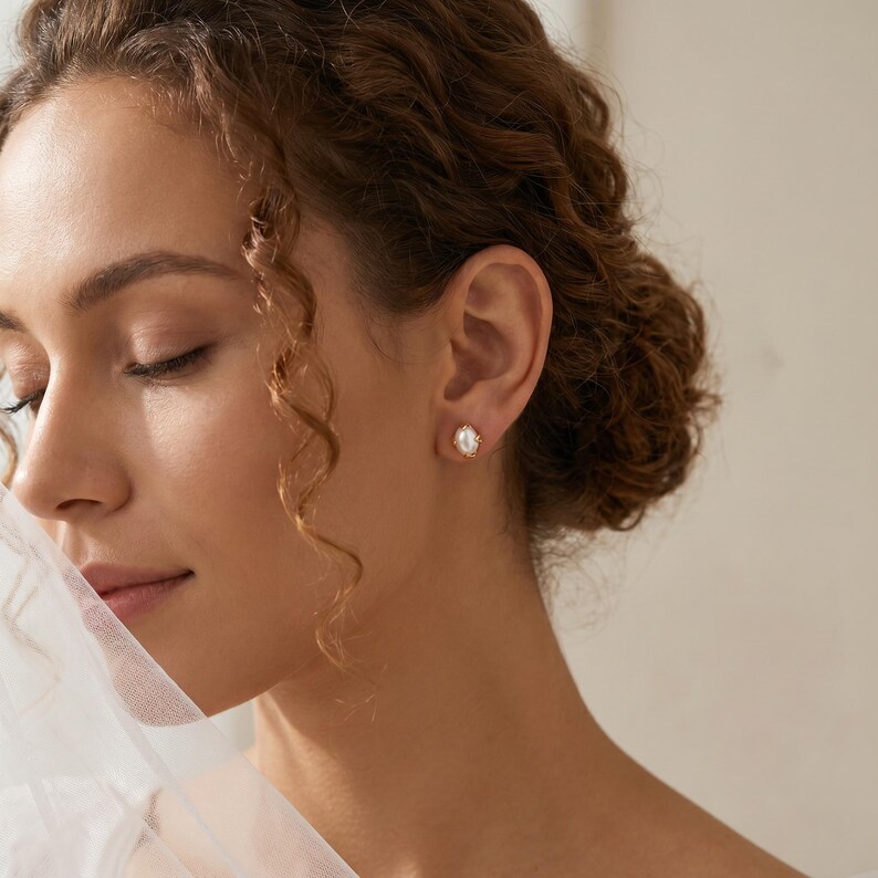 May include: A small, oval-shaped stud earring featuring a central white pearl set within a gold-toned metal frame. The earring is secured to the earlobe, showcasing a polished finish and a classic, symmetrical design that highlights the pearl.