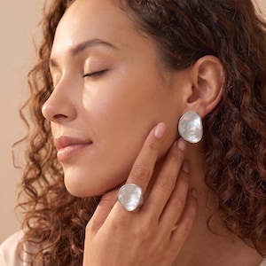 Peut inclure: Une bague et une boucle d'oreille assortie de couleur argentée, chacune dotée d'une incrustation en nacre blanche irisée de forme organique irrégulière. Les bijoux présentent un bord en métal poli lisse encadrant la surface nacrée scintillante.
