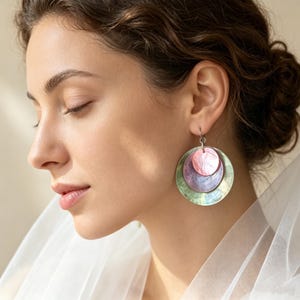Könnte beinhalten: Ein Paar Ohrhänger mit drei übereinanderliegenden, kreisförmigen Scheiben aus irisierendem Muschelmaterial. Die kleinste obere Scheibe ist rosa, die mittlere Scheibe ist hellviolett und die größte untere Scheibe ist schimmernd hellgrün, alle verbunden durch einen silbernen Haken.