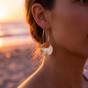 Peut inclure: Une boucle d'oreille créole en or composée d'une petite perle ronde orange et d'un pendentif plus grand en forme de disque irisé en nacre. Le bijou est suspendu à un fin anneau en fil d'or qui traverse le lobe de l'oreille.
