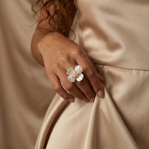 Puede incluir: Un anillo con forma de flor con pétalos blancos y un grupo de pequeñas perlas redondas en el centro. El anillo se lleva en una mano, sobre un fondo de tonos neutros.