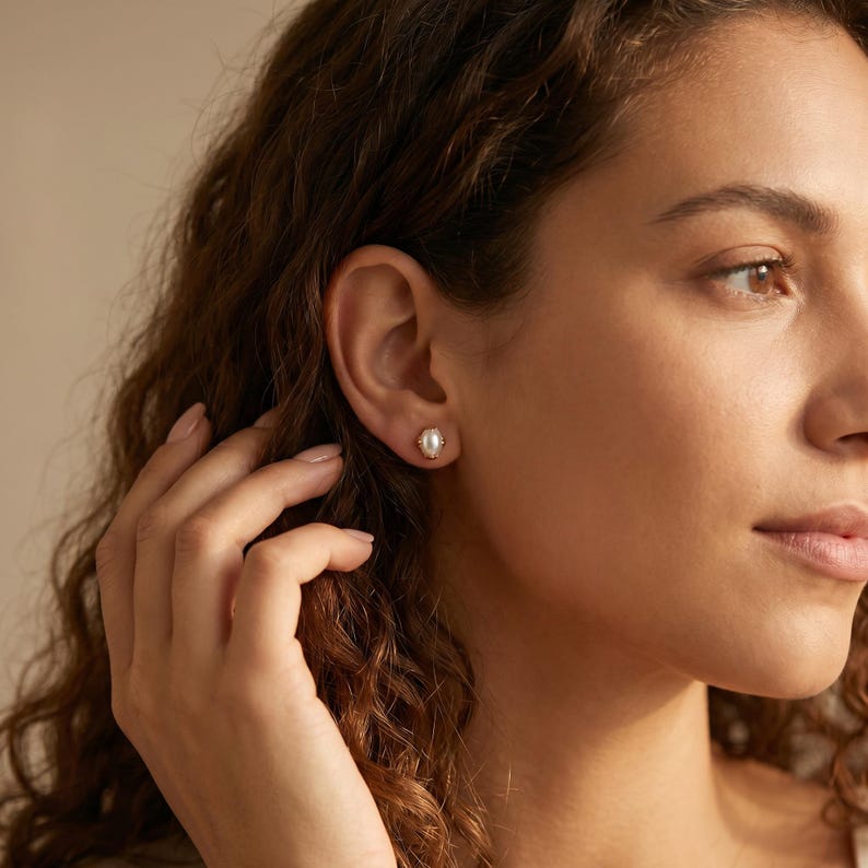 May include: A small, oval-shaped stud earring featuring a central white pearl set within a polished gold-toned metal frame. The earring is secured to the earlobe, showcasing a classic design with a smooth, lustrous finish and a secure backing.