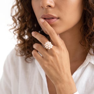 Può includere: Un anello in oro con un gruppo di piccole perle bianche rotonde disposte a forma sferica. L'anello è indossato su un dito, mettendo in risalto la fascia in oro lucido e la densa montatura di perle che crea un punto focale arrotondato.