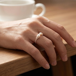 Puede incluir: Un anillo con dos pequeñas perlas blancas de forma ovalada montadas sobre una fina banda de alambre de color dorado. El alambre está enrollado alrededor de la base de cada perla, asegurándolas sobre la delicada banda metálica que rodea el dedo.