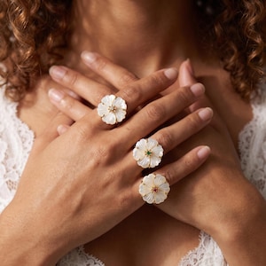 Op de afbeelding: Drie gouden ringen met witte parelmoeren bloemblaadjes. De bovenste ring heeft een heldere middensteen, de middelste ring een groene middensteen en de onderste ring een rode middensteen. Elke ring is gezet op een dunne gouden band.