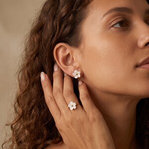 May include: A set of jewelry consisting of a stud earring and a ring, both featuring a white, five-petaled flower design with a small gold-toned center. The flower petals have a pearlescent, iridescent finish, and the metal bands are gold-toned.