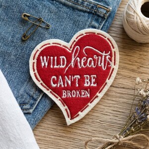 Puede incluir: Parche rojo en forma de corazón con texto bordado en blanco: "WILD hearts CAN'T BE BROKEN". El parche está sobre unos vaqueros azules. También se ve un imperdible dorado y una bobina de hilo blanco.