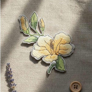 Op de afbeelding: Geborduurde bloemenpatch met een gele hibiscusbloem met groene bladeren en knoppen. De patch heeft een witte rand en is geplaatst tegen een neutrale linnen achtergrond. Een takje lavendel en een knoop zijn ook zichtbaar.