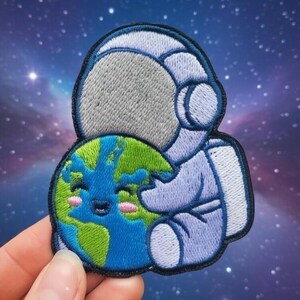 Könnte beinhalten: Gestickter Aufnäher mit einem hellblauen Astronauten, der die lächelnde Erde umarmt. Die Erde ist mit grünen Kontinenten, blauen Ozeanen und rosigen Wangen dargestellt. Der Aufnäher hat einen schwarzen Rand und einen Weltraumhintergrund.