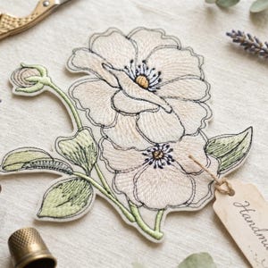 Peut inclure: Écusson floral brodé avec deux grandes fleurs crème, des contours noirs et des centres jaunes. Comprend des feuilles vertes et un bouton floral. Une petite étiquette avec "Handmade" est attachée. Le patch est sur un tissu crème.