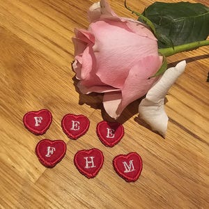 May include: A pale pink rose with a green stem and leaf rests on a wooden surface. Six small red heart-shaped patches with embroidered white letters are scattered around the rose. The letters are F, E, E, F, H, and M.