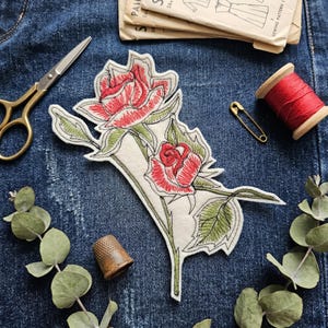 Könnte beinhalten: Ein gestickter Rosenaufnäher mit roten Blüten und grünen Blättern auf cremefarbenem Hintergrund. Nähwerkzeuge wie goldene Schere, rote Garnrolle und Fingerhut sind auf einem Jeansstoff arrangiert, was das Thema Handwerk unterstreicht.