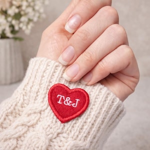 May include: A red heart-shaped patch embroidered with the letters "T&J" in white thread, attached to a cream cable-knit sweater cuff. The image shows a close-up of the sweater and a hand.