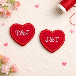 Może przedstawiać: Dwie czerwone naszywki w kształcie serca z białymi haftowanymi inicjałami, "T&J" i "J&T". Naszywki mają czerwoną, szytą obwódkę. Obraz zawiera różowe konfetti w kształcie serca, różowe kwiaty i szpulkę czerwonej nici.