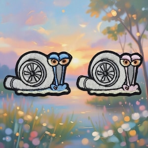 May include: Two embroidered patches featuring a snail design with a turbocharger shell. The snails have large, cartoonish eyes and are in light blue and pink colors. The patches have a black border and are set against a colorful background.