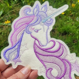 Puede incluir: Parche de unicornio bordado en tonos morado, lavanda y plateado. El unicornio tiene un cuerpo blanco, una melena fluida y un cuerno brillante. El parche está hecho de fieltro y presenta intrincados detalles de costura, ideal para ropa o accesorios.