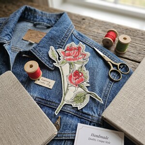 Puede incluir: Chaqueta vaquera azul con un parche bordado de rosas rojas y verdes. La imagen incluye carretes de hilo, tijeras y una etiqueta que dice "Rosemary Crafts - Etsy". Un letrero dice "Handmade Quality, Unique Style".