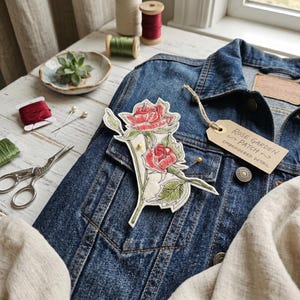 Op de afbeelding: Een blauw spijkerjack met een bloemen geborduurde patch met rode rozen en groene bladeren. De patch is vastgezet met gouden spelden. Een label leest "Rose Garden Patch, Embroidered Detail."