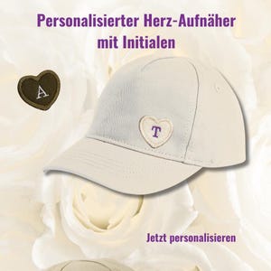 May include: A light beige baseball cap with a heart-shaped patch embroidered with the letter "T". A separate heart-shaped patch with the letter "A" is also visible. The text "Personalisierter Herz-Aufnäher mit Initialen" and "Jetzt personalisieren" are displayed.