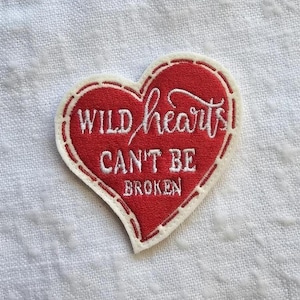 Könnte beinhalten: Ein herzförmiger, gestickter Aufnäher mit roter Mitte und weißem Rand. Der Aufnäher trägt die Worte "WILD hearts CAN'T BE BROKEN" in weißer Schrift und Blockbuchstaben.