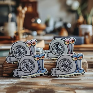 May include: Four embroidered patches featuring a cartoon snail with a turbocharger shell. The snails have gray shells, blue and pink eye stalks, and yellow eyes. The patches are on a wooden surface.
