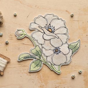 Könnte beinhalten: Gestickter Blumenaufnäher mit zwei großen, cremefarbenen Blüten mit detaillierten Stichen und einer Knospe. Der Aufnäher hat grüne Blätter und Stiele mit einem cremefarbenen Rand. Das Design befindet sich auf einer hellen Holzoberfläche.