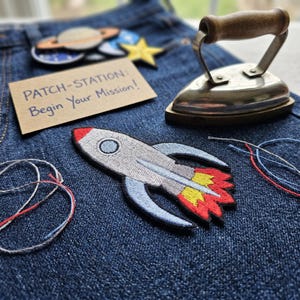 Könnte beinhalten: Eine Sammlung von Weltraum-Bügelbildern, darunter eine Rakete, ein Planet und ein Stern, auf Denim-Stoff angeordnet. Ein Vintage-Bügeleisen und eine Karte mit der Aufschrift "PATCH-STATION: Begin Your Mission!" sind ebenfalls vorhanden.