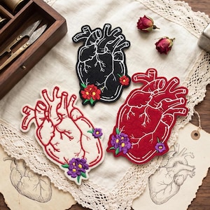 May include: Three embroidered heart patches in black, red, and off-white, each with white stitching and floral accents. The patches are arranged on a lace-trimmed cloth, with dried roses and vintage-style tags nearby. The patches are approximately 10 cm in height.